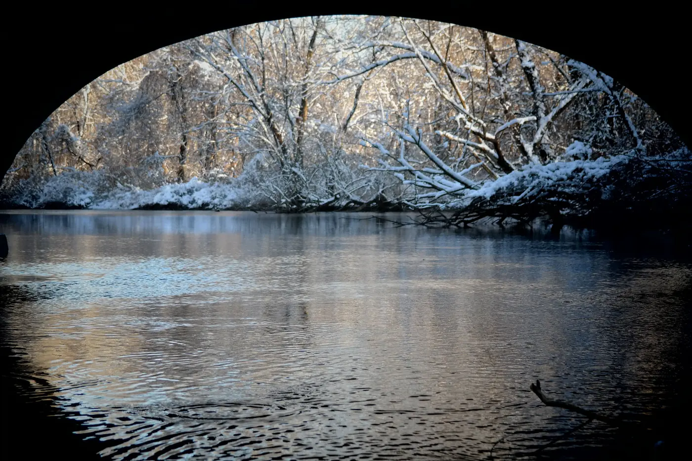 The view from beneath a bridge arch across a snow-lined winter river.